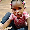 toddler, child, baby, portrait, face, looking_at_camera, curious, serious, hair_beads, braids, pink_clothing, polka_dots, short_sleeves, sitting, indoor, wooden_floor, toy, adult_lap, close_up, photograph