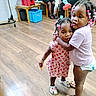 children, siblings, girls, toddler, preschooler, hugging, portrait, smiling, indoors, classroom, wooden_floor, chairs, storage_bin, colorful_hair_beads, braids, pink_clothing, dress, diaper, clogs, candid_photo