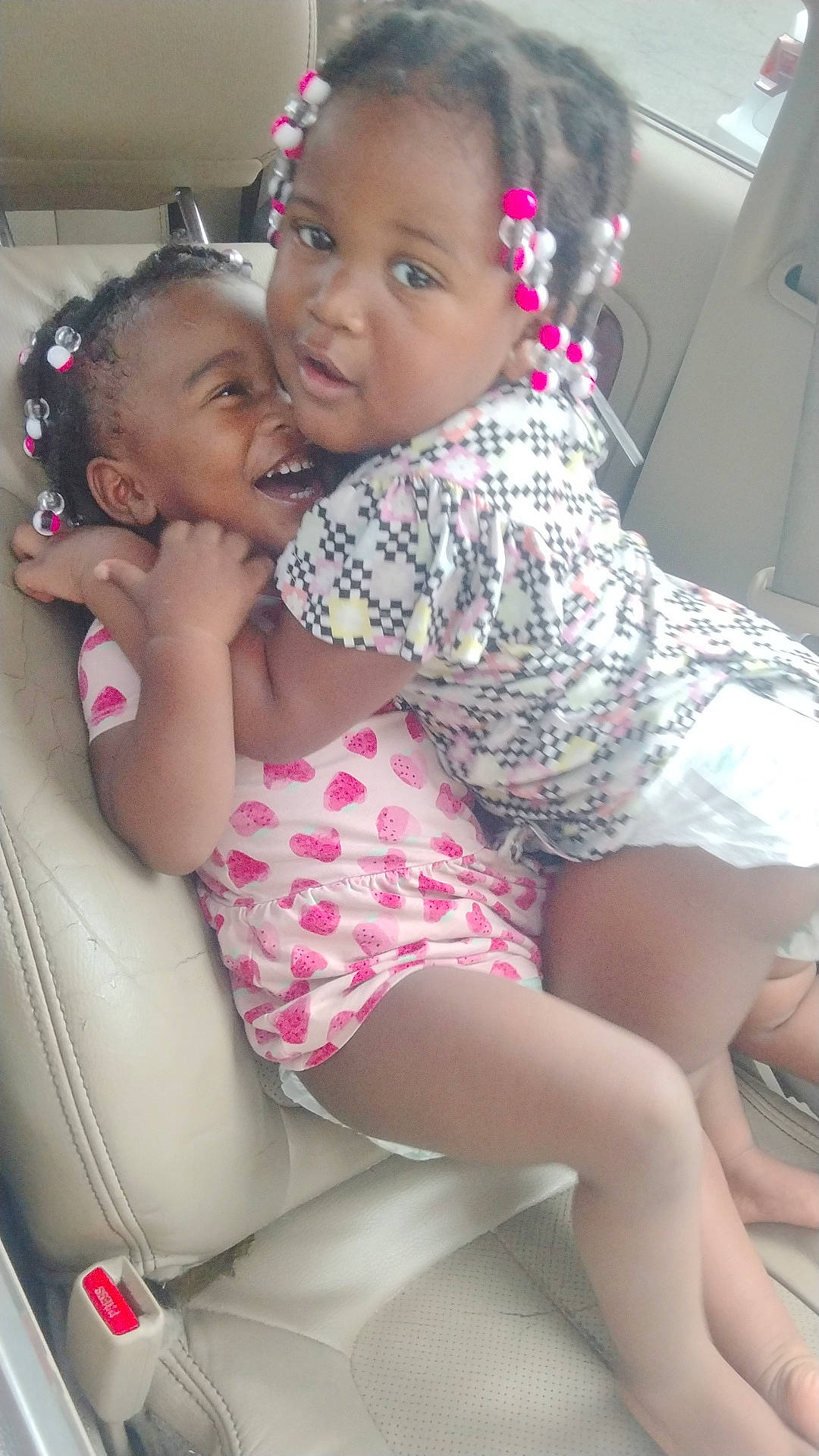 Alaia is registered to the contest to win money with this photo:  smiling, beaded_hair, braids, car, car_seat, children, colorful_clothing, cuddling, diaper, dress, family, girls, happy, laughing, playful, portrait, siblings, sitting, toddler, vehicle_interior