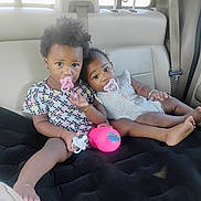 Alaia is registered to the contest to win money with this photo: children, toddlers, babies, siblings, car, backseat, pacifiers, toy, pink_toy, curly_hair, smiling, sitting, lying_down, infants, dress, floral_clothing, bracelet, vehicle_interior, road_trip, portrait