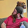 man, dog, beard, glasses, cap, couch, indoor, lotion_bottle, wall, framed_photo, red_shirt, pet, shoulder, seated, relaxed, light_switch, brown_dog, casual, home, looking_away