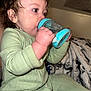 toddler, child, baby, onesie, green_clothing, sippy_cup, curly_hair, drinking, indoor, blanket, closeup, cute, portrait, hand, face, sitting, soft_lighting, cozy, young_child, focused