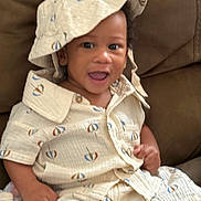 Jeremiah is registered to the contest to win money with this photo: toddler, child, baby, smiling, hat, bucket_hat, shirt, shorts, clothing, indoors, couch, furniture, toy, colorful_toy, happy, face, person, sitting, cute, playful