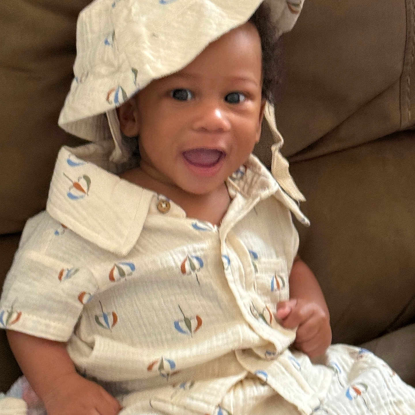 Jeremiah is registered to the contest to win money with this photo: toddler, child, baby, smiling, hat, bucket_hat, shirt, shorts, clothing, indoors, couch, furniture, toy, colorful_toy, happy, face, person, sitting, cute, playful