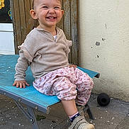 Alva participe au concours pour gagner de l'argent avec cette photo : toddler, child, smiling, sitting, outdoor, clothing, shoes, chair, wooden_door, wall, happy, person, face, footwear, pavement, casual_wear, baby, joy, cute, playful