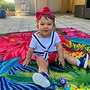 Amalya participe au concours pour gagner de l'argent avec cette photo : baby, blanket, casual_clothing, child, colorful, door, floor, flower_pattern, happy, headband, necklace, outdoor, patio, person, playful, sitting, smiling, sneakers, sunlight, toy