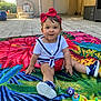 baby, blanket, casual_clothing, child, colorful, door, floor, flower_pattern, happy, headband, necklace, outdoor, patio, person, playful, sitting, smiling, sneakers, sunlight, toy