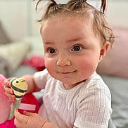 Lucy is registered to the contest to win money with this photo: baby, child, face, pigtails, toy, white_shirt, smile, indoor, cute, skin, hand, person, infant, closeup, expression, soft_light, cheeks, curly_hair, happy, playing