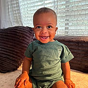 Jovon is registered to the contest to win money with this photo: baby, child, smile, happy, indoor, sitting, toddler, clothing, green_shirt, face, person, window, curtain, natural_light, cushion, soft_texture, home, portrait, cute, hands