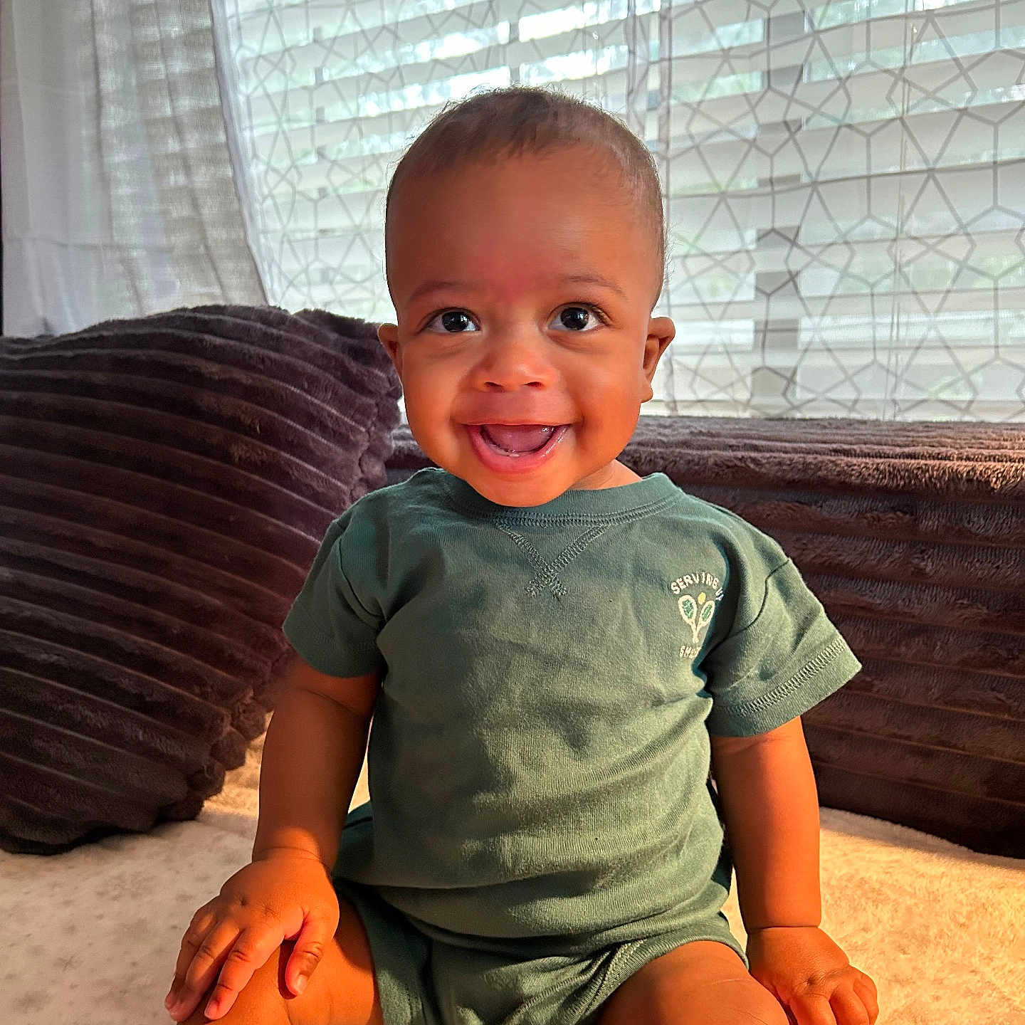 Jovon is registered to the contest to win money with this photo: baby, child, smile, happy, indoor, sitting, toddler, clothing, green_shirt, face, person, window, curtain, natural_light, cushion, soft_texture, home, portrait, cute, hands