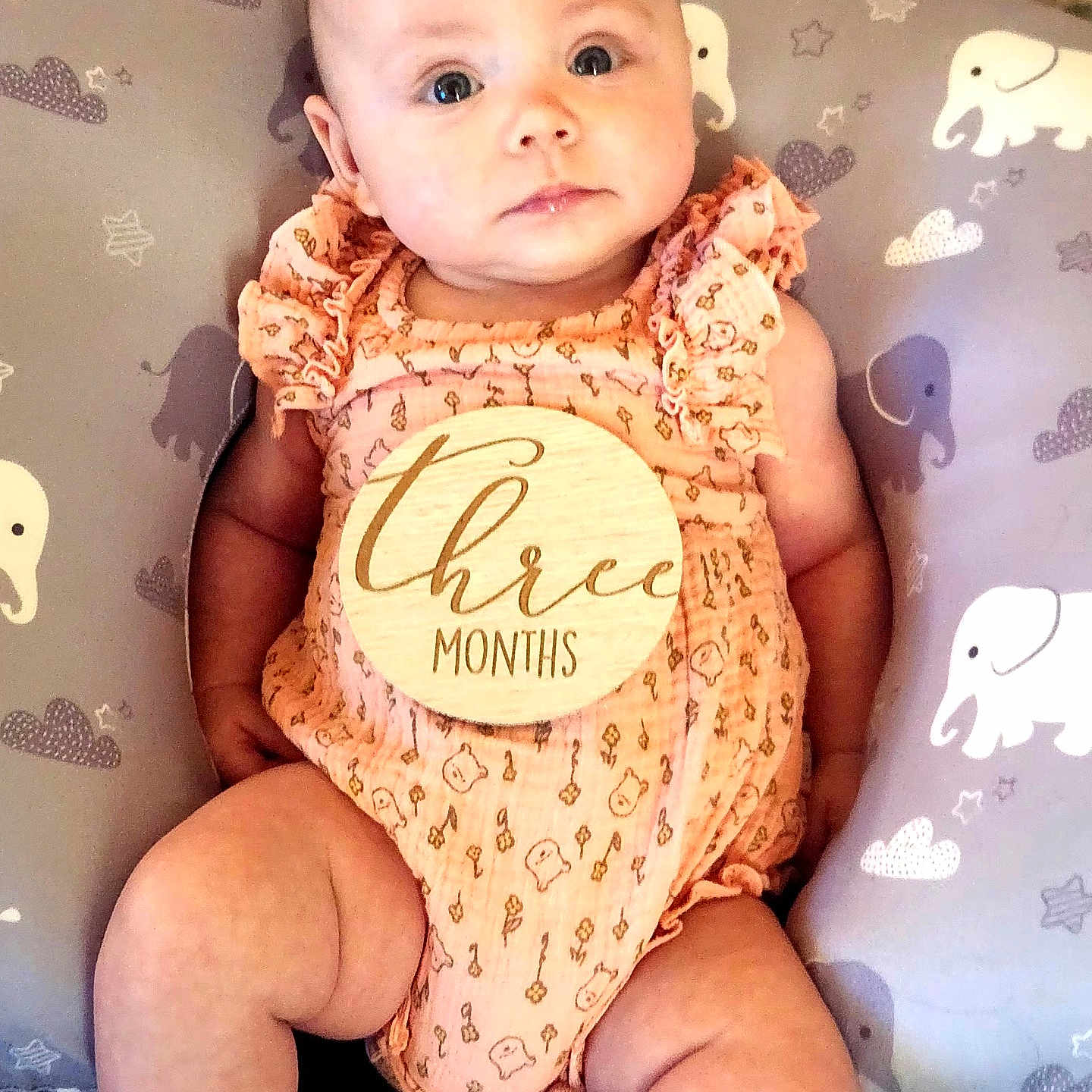 Illyana is registered to the contest to win money with this photo: baby, infant, child, onesie, peach_color, milestone, wooden_sign, three_months, pillow, elephant_pattern, cushion, cute, portrait, lying_down, skin, face, arms, legs, indoor, calm