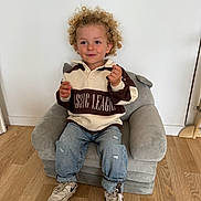 Qays a rejoint le concours — aidez-le/la à gagner de superbes lots ! toddler, child, curly_hair, sweater, jeans, sneakers, chair, plush_chair, wooden_floor, indoor, smiling, casual_clothing, white_wall, footwear, small_chair, cute, person, young_child, seated, happy