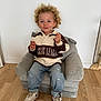 toddler, child, curly_hair, sweater, jeans, sneakers, chair, plush_chair, wooden_floor, indoor, smiling, casual_clothing, white_wall, footwear, small_chair, cute, person, young_child, seated, happy
