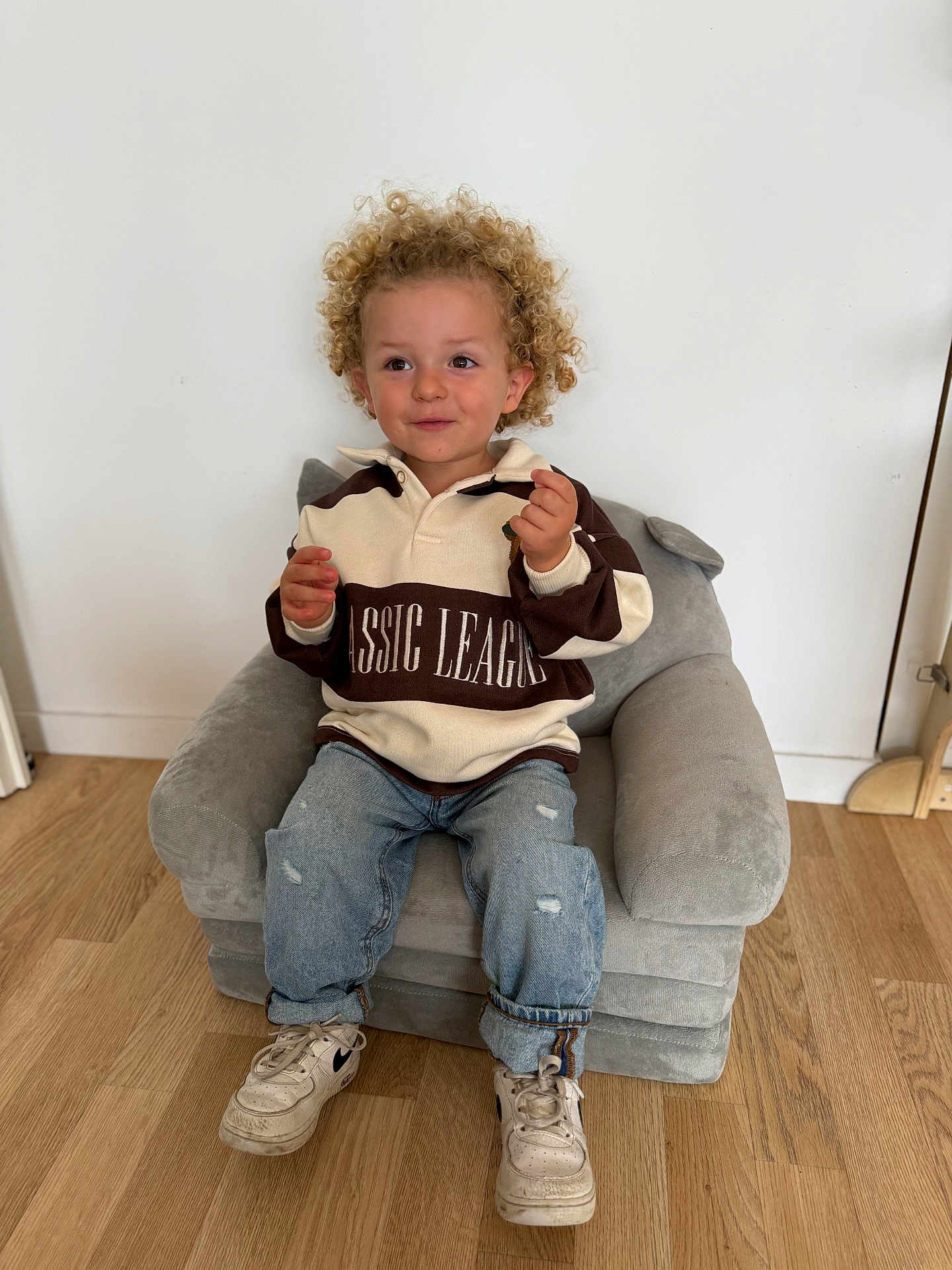 Qays a rejoint le concours — aidez-le/la à gagner de superbes lots ! toddler, child, curly_hair, sweater, jeans, sneakers, chair, plush_chair, wooden_floor, indoor, smiling, casual_clothing, white_wall, footwear, small_chair, cute, person, young_child, seated, happy