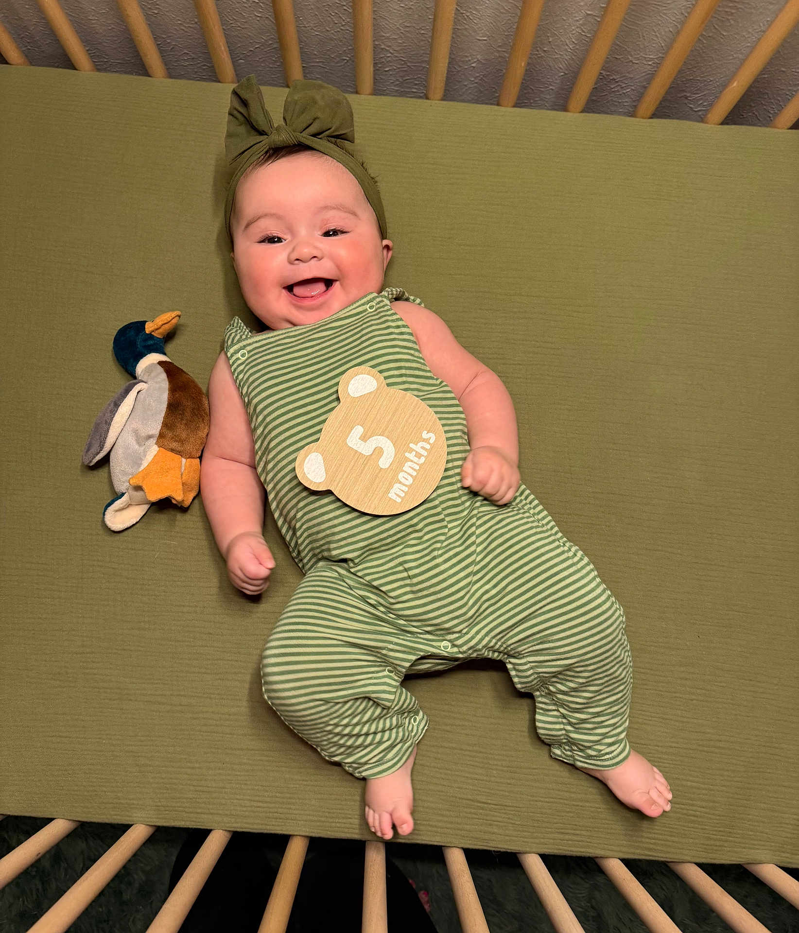 Tatum is registered to the contest to win money with this photo: baby, infant, smile, crib, crib_slats, headband, bow, onesie, striped_outfit, plush_toy, duck, milestone_sign, green_sheet, hands, feet, portrait, happy, cute, lying_down, nursery