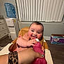baby, smiling_baby, high_chair, pink_bottle, adult_hand, leopard_scrunchie, checkered_nails, tongue_out, rosy_cheeks, indoor, window_blinds, water_dispenser, cardboard_box, countertop, wood_floor, feeding, portrait, happy, playful, hand_grip