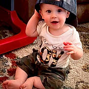 Karter is registered to the contest to win money with this photo: baby, child, cap, hat, sitting, carpet, indoor, camouflage, shorts, shirt, expression, cute, playful, person, foot, hand, smile, bright_eyes, home, infant