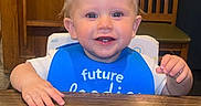 Karter is registered to the contest to win money with this photo: baby, child, toddler, blue_bib, smiling, table, wooden_table, wooden_chair, indoor, face, person, cute, happy, blond_hair, blue_eyes, sitting, highchair, clothing, feeding, home