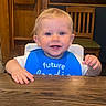 baby, child, toddler, blue_bib, smiling, table, wooden_table, wooden_chair, indoor, face, person, cute, happy, blond_hair, blue_eyes, sitting, highchair, clothing, feeding, home