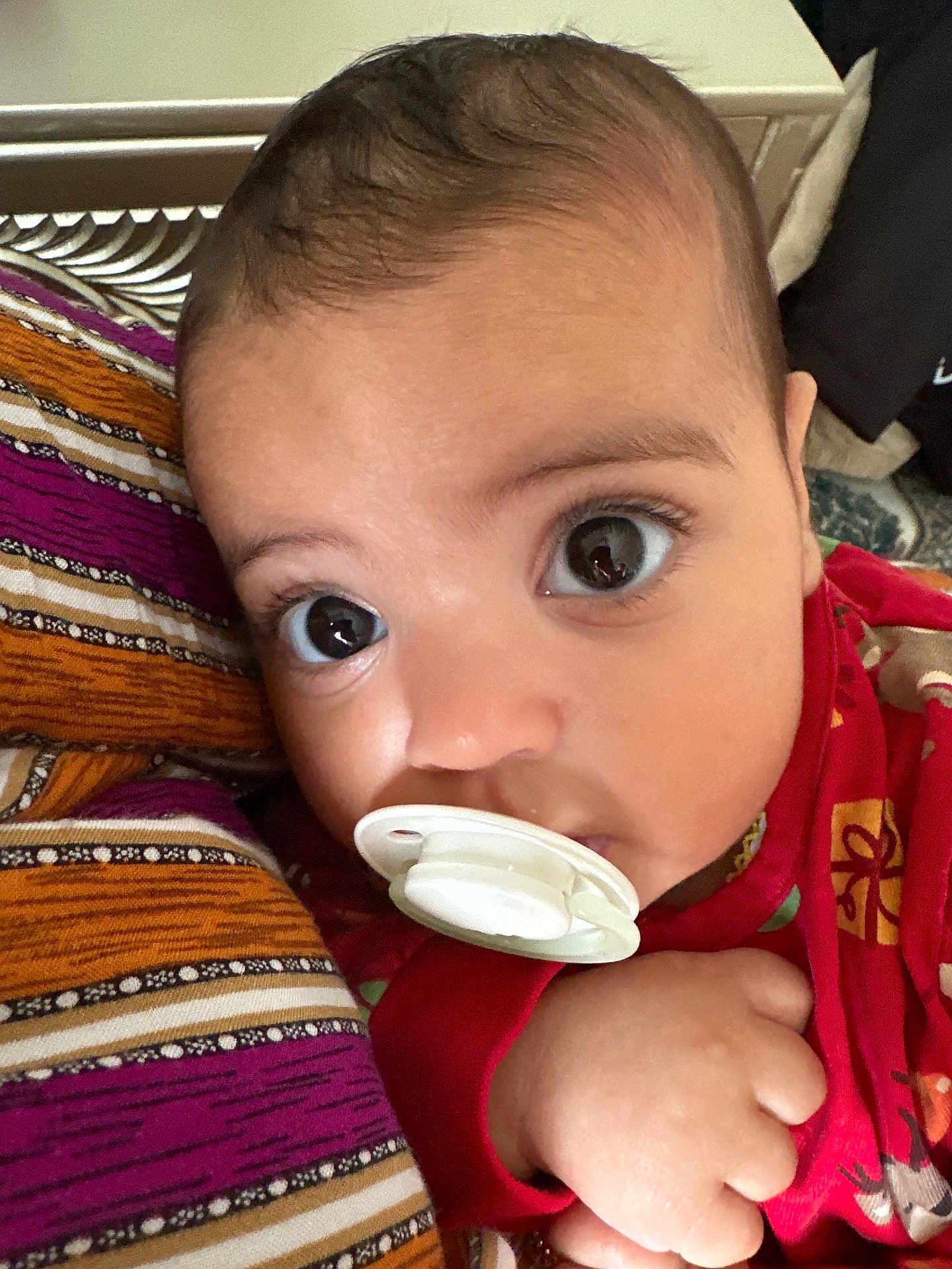Inaya Rekad a rejoint le concours — aidez-le/la à gagner de superbes lots ! baby, pacifier, close_up, face, hand, curious, wide_eyes, patterned_fabric, red_clothing, infant, skin, portrait, cute, child, indoors, young_child, person, baby_girl, baby_boy, sleepwear