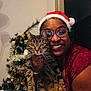 person, cat, tabby_cat, santa_hat, glasses, christmas_tree, ornaments, holiday, smile, indoor, celebration, festive, red_clothing, decorations, tree, happy, holding, pet, home, cheerful