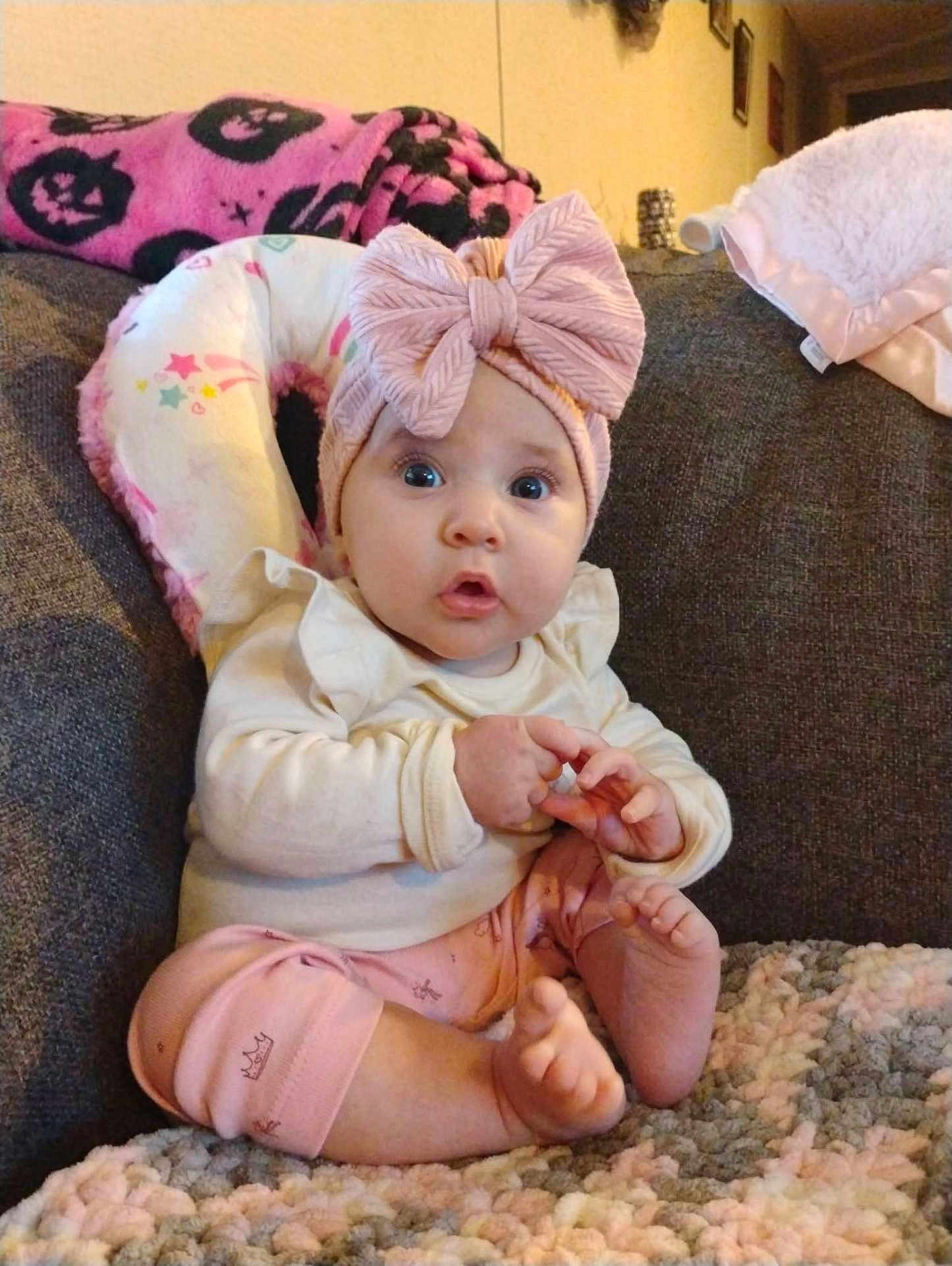 Alyana is registered to the contest to win money with this photo: baby, child, infant, pink_bow, headband, couch, blanket, clothing, sitting, indoor, cute, expression, hands, feet, person, portrait, soft_toys, cozy, curious, home