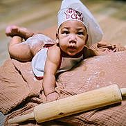 Jahdrian joined the competition — help win amazing prizes! baby, infant, chef_hat, baby_chef, rolling_pin, flour, brown_cloth, cooking_theme, cute, curious_expression, baking, child, indoors, portrait, soft_lighting, flooring, apron, hands, face, laying_down