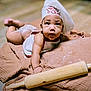 baby, infant, chef_hat, baby_chef, rolling_pin, flour, brown_cloth, cooking_theme, cute, curious_expression, baking, child, indoors, portrait, soft_lighting, flooring, apron, hands, face, laying_down