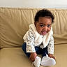 adorable, baby, child, couch, curly_hair, dark_eyes, earrings, faux_fur_jacket, floral_pattern, happy, indoor, infant, jeans, playful, portrait, sitting, smile, sofa, tongue_out, white_shoes