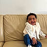 baby, beige, child, clothing, couch, curly_hair, fur_vest, happy, indoor, infant, jeans, living_room, portrait, seated, skin, smile, sneakers, sofa, tiny_shoes, toyless