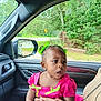 toddler, child, car_interior, pink_dress, green_bows, person, hand, window, side_view_mirror, trees, road_sign, seat, curious, expression, earrings, adult_hand, vehicle, casual, daylight, portrait