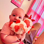 Elizabeth joined the competition — help win amazing prizes! baby, child, stuffed_toy, plush, pink_clothing, heart_pattern, indoor, curtains, wall_art, person, holding, cute, infant, soft_toy, face, legs, colorful, cozy, light, gaze