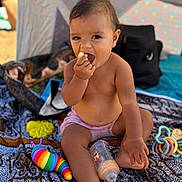 Mahana a rejoint le concours — aidez-le/la à gagner de superbes lots ! toddler, child, baby, eating, snack, bottle, toys, colorful, blanket, patterned, outdoor, sun_tent, shade, summer, bare_chest, sitting, curious, mischievous, hand, foot
