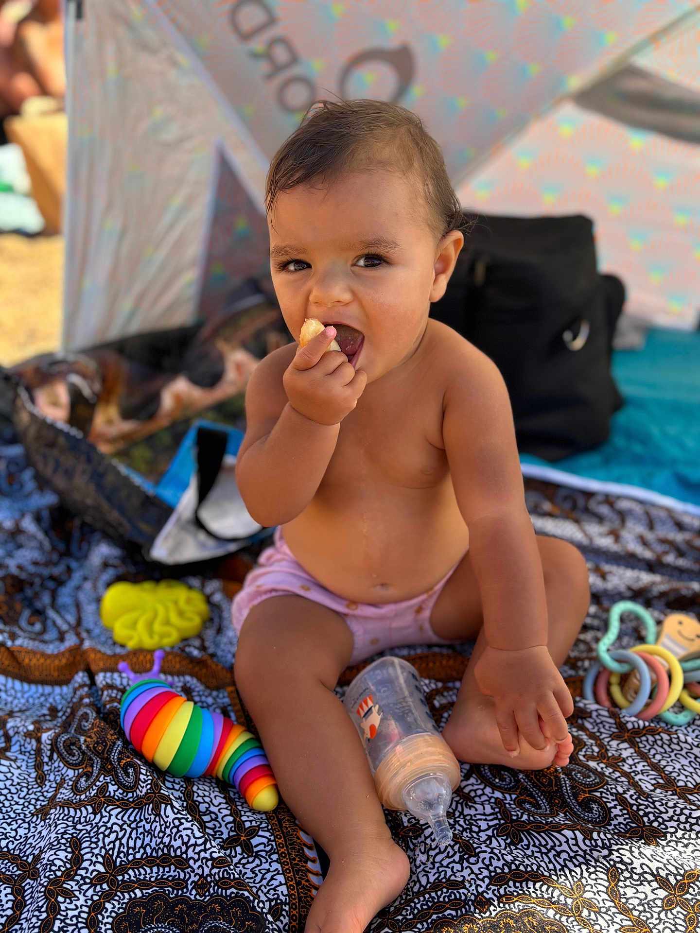 Mahana a rejoint le concours — aidez-le/la à gagner de superbes lots ! toddler, child, baby, eating, snack, bottle, toys, colorful, blanket, patterned, outdoor, sun_tent, shade, summer, bare_chest, sitting, curious, mischievous, hand, foot