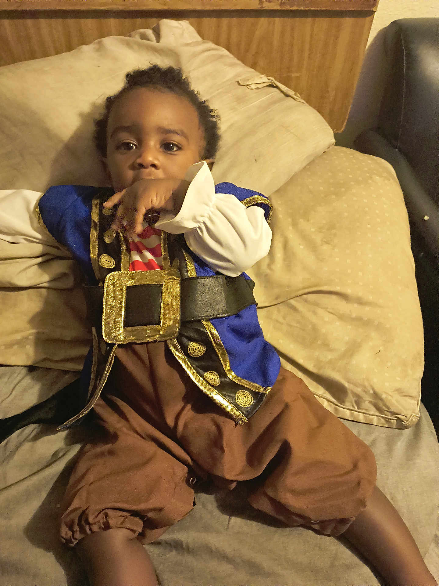 Sion is registered to the contest to win money with this photo: toddler, child, pirate_costume, blue_jacket, brown_pants, bed, pillows, hand, face, curious, indoor, young_child, sitting, costume, clothing, person, cute, portrait, resting, infant