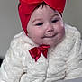 baby, child, face, headband, red_bow, white_coat, portrait, cute, infant, smile, soft_lighting, warm_clothing, indoor, closeup, adorable, young_child, expression, fashion_accessory, headwear, skin