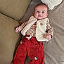baby, infant, smiling, red_pants, striped_shirt, vest, couch, cushion, happy, face, person, child, lying_down, cozy, soft_texture, indoor, cute, clothing, small_hand, bright_eyes