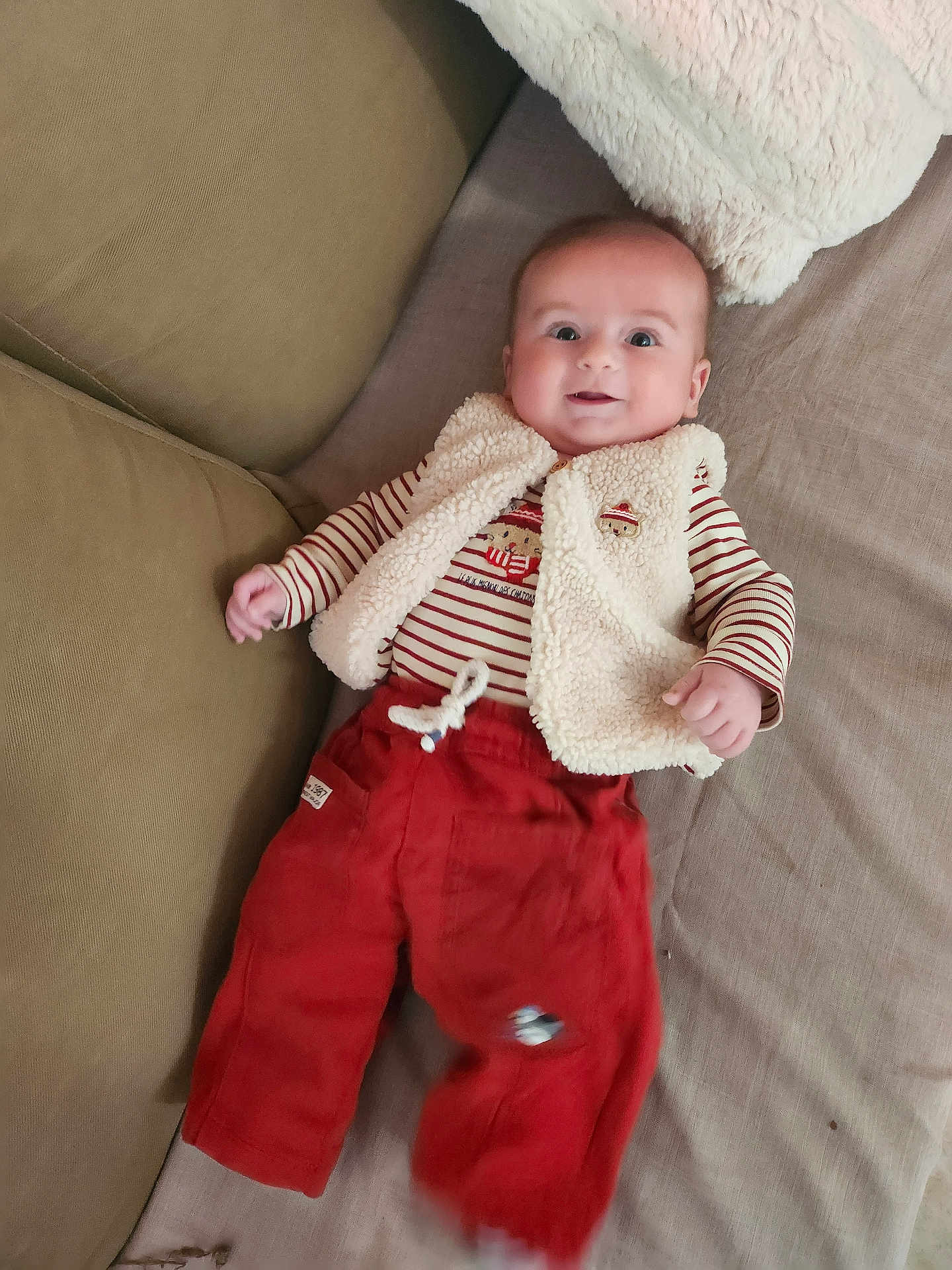 Noam a rejoint le concours — aidez-le/la à gagner de superbes lots ! baby, infant, smiling, red_pants, striped_shirt, vest, couch, cushion, happy, face, person, child, lying_down, cozy, soft_texture, indoor, cute, clothing, small_hand, bright_eyes