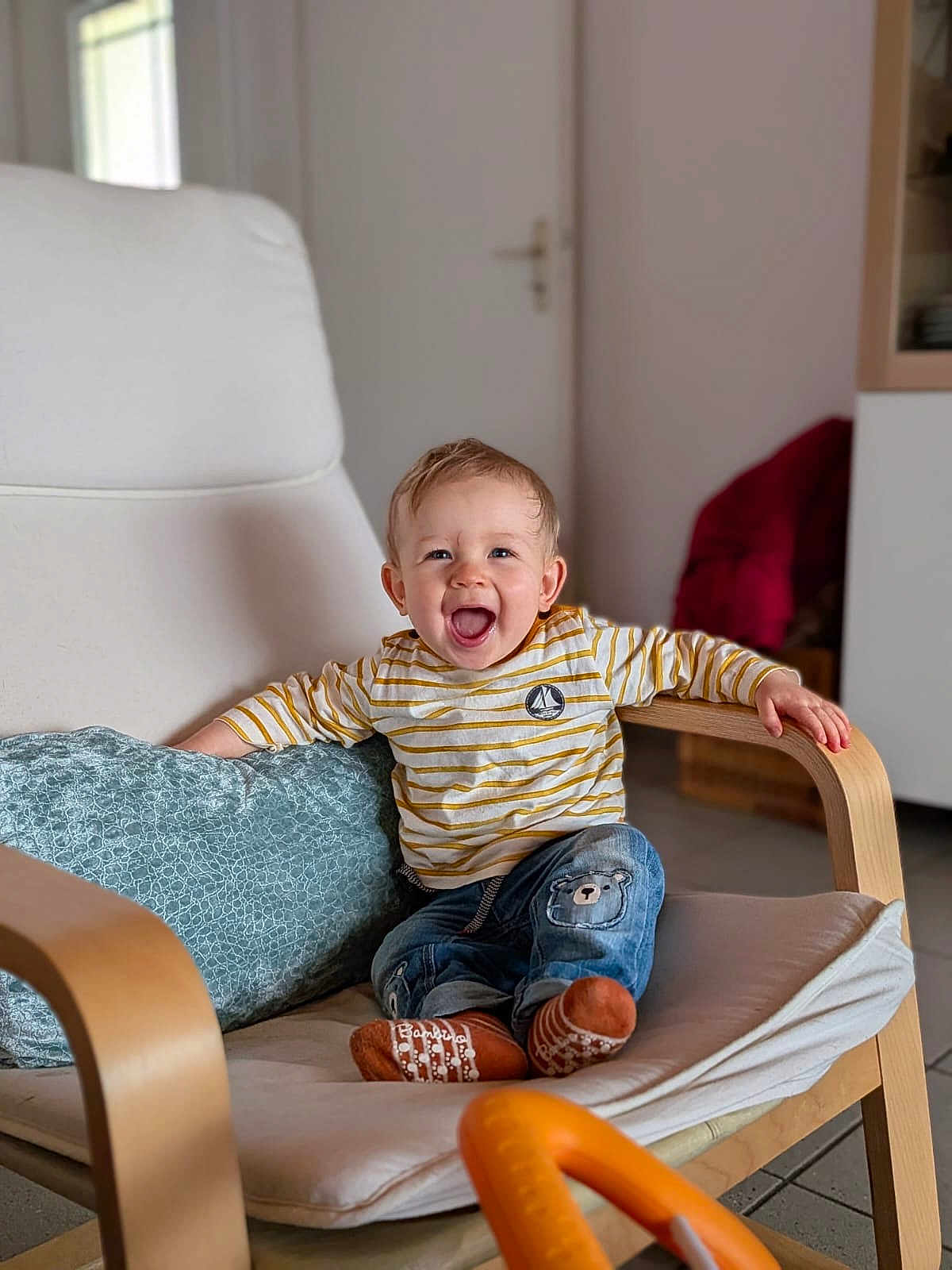 Maël a rejoint le concours — aidez-le/la à gagner de superbes lots ! toddler, child, smiling, happy, indoor, chair, cushion, wooden_furniture, striped_shirt, jeans, socks, face, person, home, living_room, baby, cute, joy, seated, casual_clothing