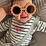 baby, child, sunglasses, striped_clothing, smile, happy, infant, face, hand, quilted_cushion, text_on_clothing, red_text, fabric, seat_belt, cute, indoor, person, head, closeup, playful