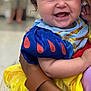 baby, child, smile, happy, curly_hair, red_bow, blue_dress, yellow_skirt, lace_sock, costume, portrait, holding, adult, arm, indoor, blurred_background, person, cute, celebration, closeup