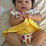 baby, child, infant, curly_hair, bow, blanket, yellow_cloth, fruit_pattern, onesie, hands, feet, lying_down, cute, chubby_cheeks, looking_at_camera, indoor, soft_lighting, skin, portrait, adorable