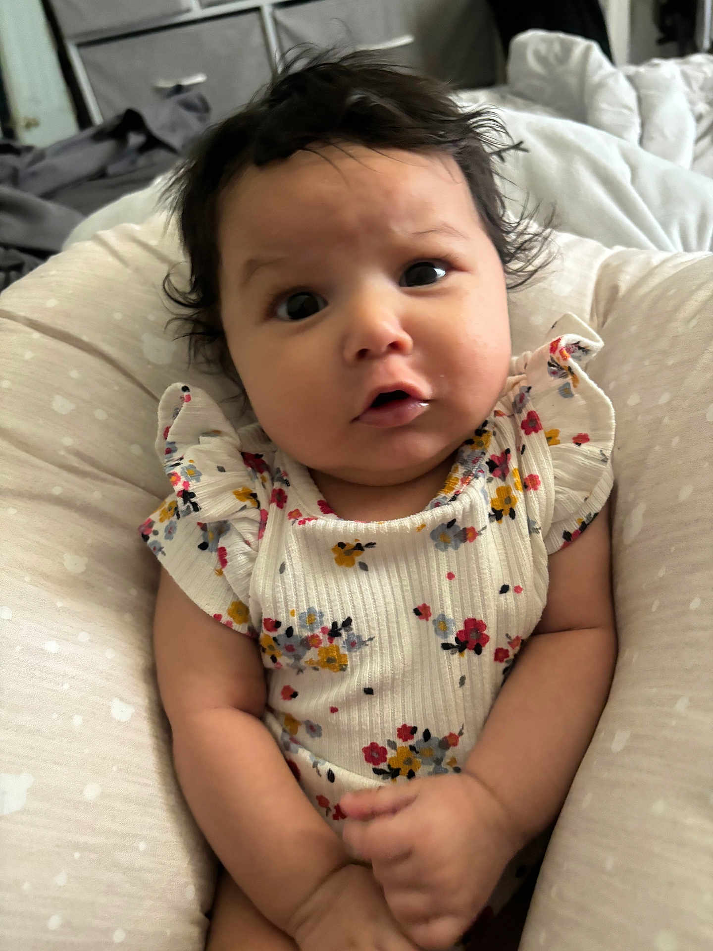 Anivia is registered to the contest to win money with this photo: baby, infant, child, face, eyes, hair, clothing, floral_pattern, sitting, cushion, indoor, soft_texture, cute, expression, portrait, skin, hand, person, young, comfort