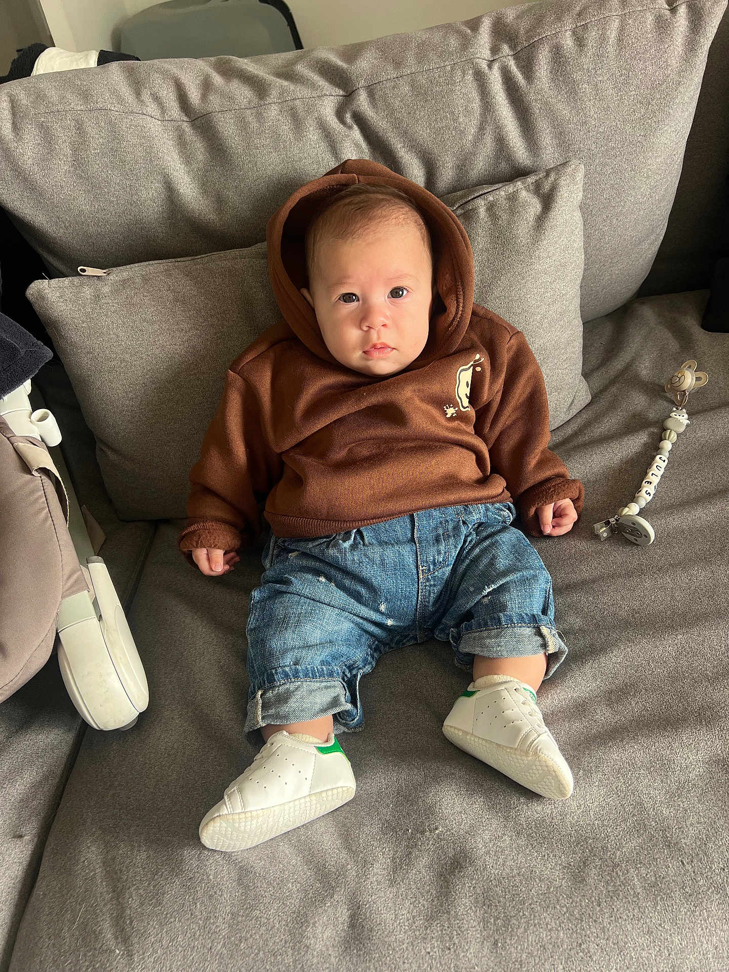 Jules a rejoint le concours — aidez-le/la à gagner de superbes lots ! baby, child, hoodie, denim, jeans, sneakers, white_shoes, couch, gray_couch, pillow, pacifier_clip, indoor, cute, infant, sitting, face, person, clothing, footwear, relaxed