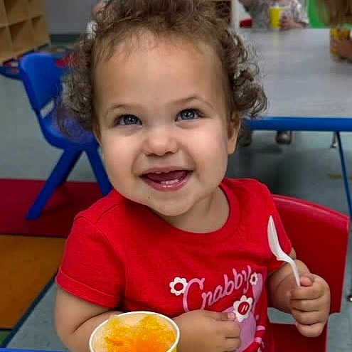 Emerson is registered to the contest to win money with this photo: toddler, child, smiling, curly_hair, red_shirt, spoon, yellow_cup, table, indoor, daycare, children, happy, playroom, blue_chair, food, snack, person, portrait, cute, seated