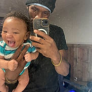 Zamir is registered to the contest to win money with this photo: baby, child, man, mirror, selfie, phone, beanie, black_shirt, striped_onesie, laughing, smiling, hand, holding, reflection, indoor, wall, bracelet, plaid_pants, person, happy