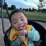 baby, child, swing, playground, outdoor, park, smiling, tongue_out, yellow_bib, green_clothing, trees, sky, dusk, blurred_background, happy, person, infant, seat, play, nature