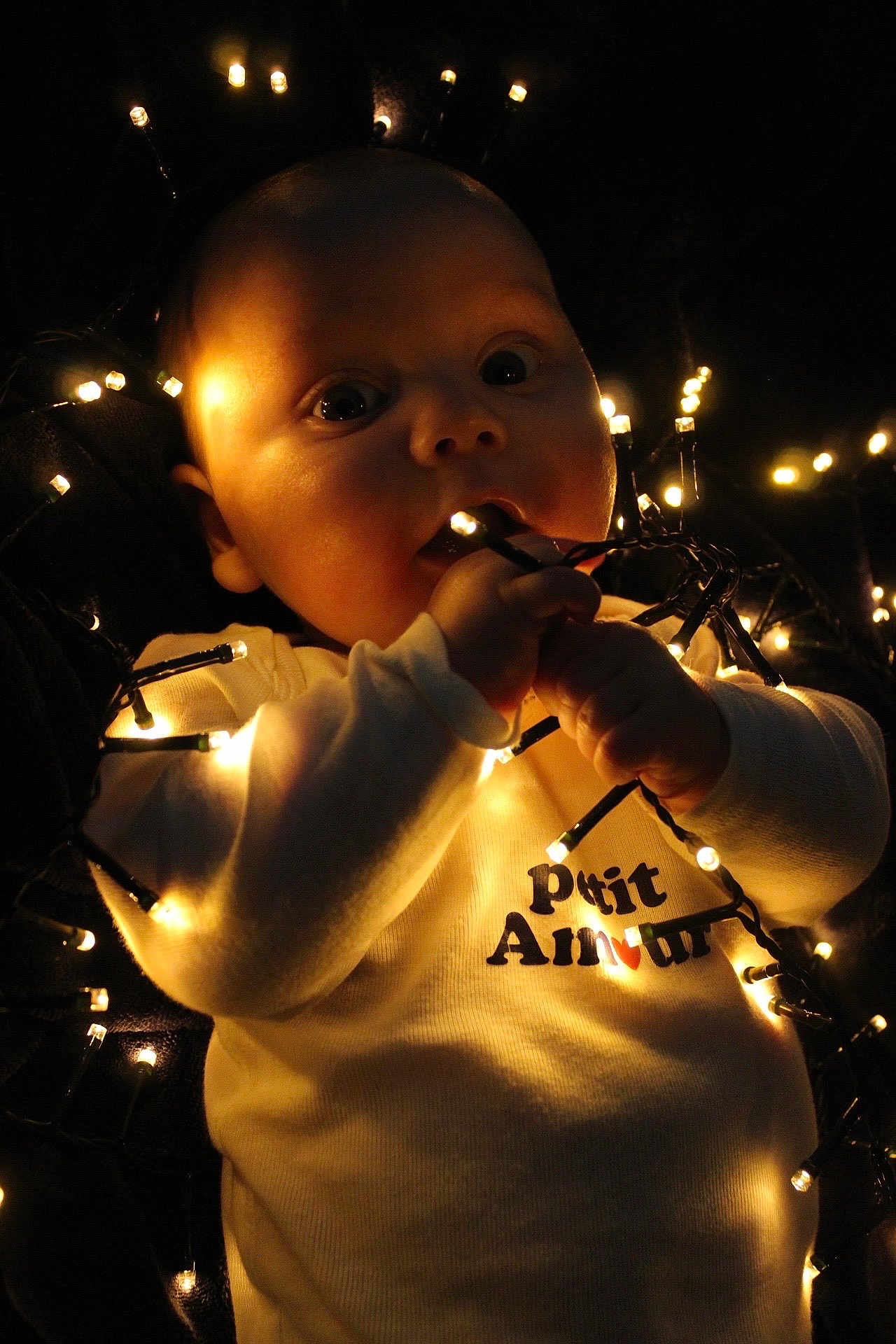Elyo participe au concours pour gagner de l'argent avec cette photo : baby, infant, child, lights, string_lights, glowing, dark_background, face, hands, chewing, curious, white_clothing, shirt, text, ptit_amour, closeup, portrait, warm_light, indoors, cute