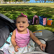 Anaya is registered to the contest to win money with this photo: baby, child, stroller, pink_top, floral_skirt, headband, outdoor, park, grass, trees, sunlight, happy, smiling, person, infant, daylight, nature, jar, bags, seated