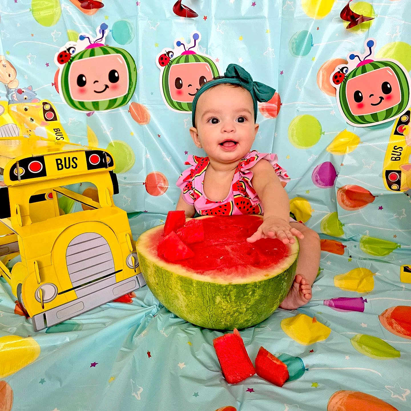 Astrid is registered to the contest to win money with this photo: baby, bright_colors, child, co_comelon, colorful_background, cute, dress, fruit, fruit_pieces, happy, headband, indoors, infant, party_decorations, playful, ribbons, sitting, smiling, toy_bus, watermelon
